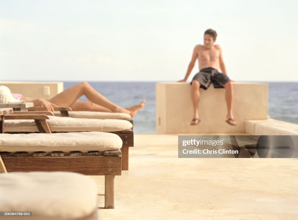 Woman sunbathing on pool terrace, man sitting on wall in background