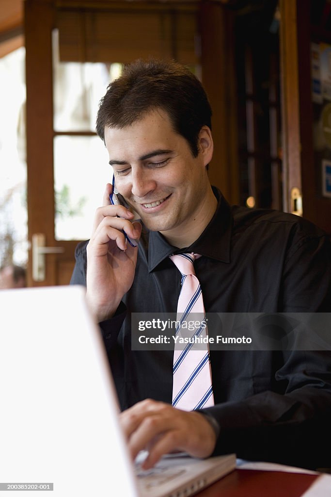 Businessman working on laptop, talking on mobile phone, smiling