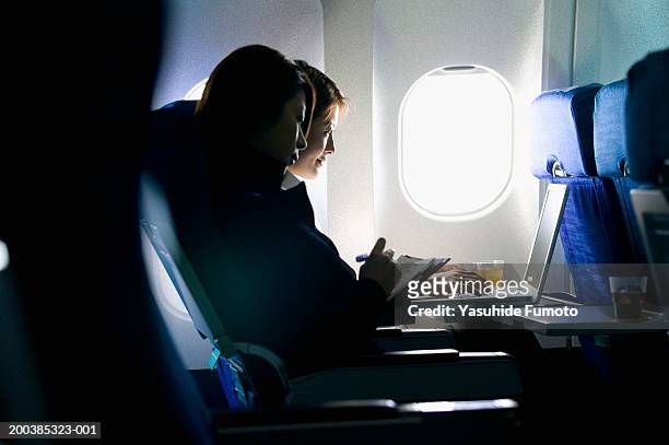 two young businesswomen sitting in airplane, working on laptop - raamplaats stockfoto's en -beelden
