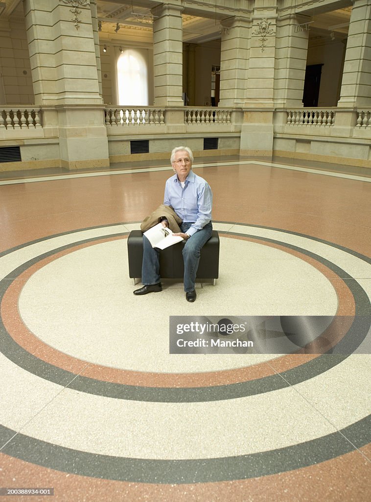 Senior man sitting on bench in museum concourse, holding book