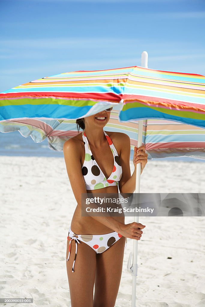 Young woman standing under beach umbrella, smiling, close-up