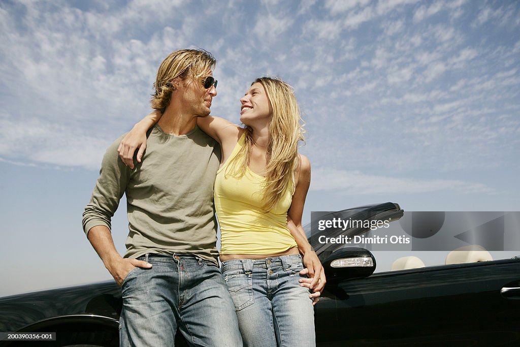Young couple leaning against car, arms around each other, smiling