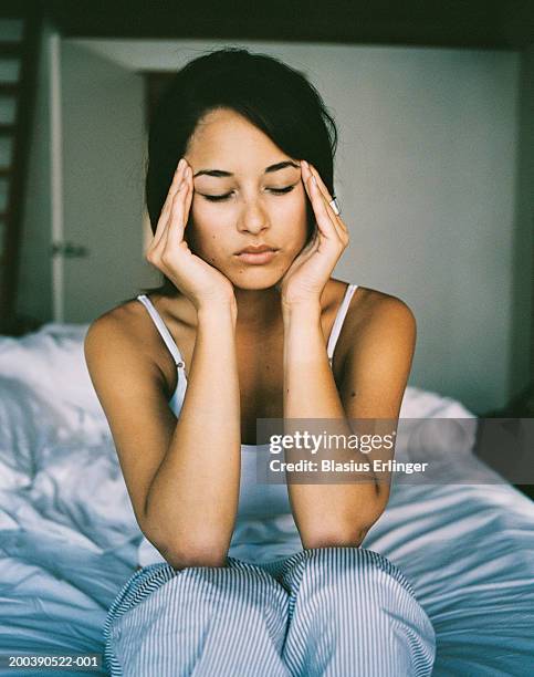 young woman sitting at edge of bed, touching temples - headache stock-fotos und bilder