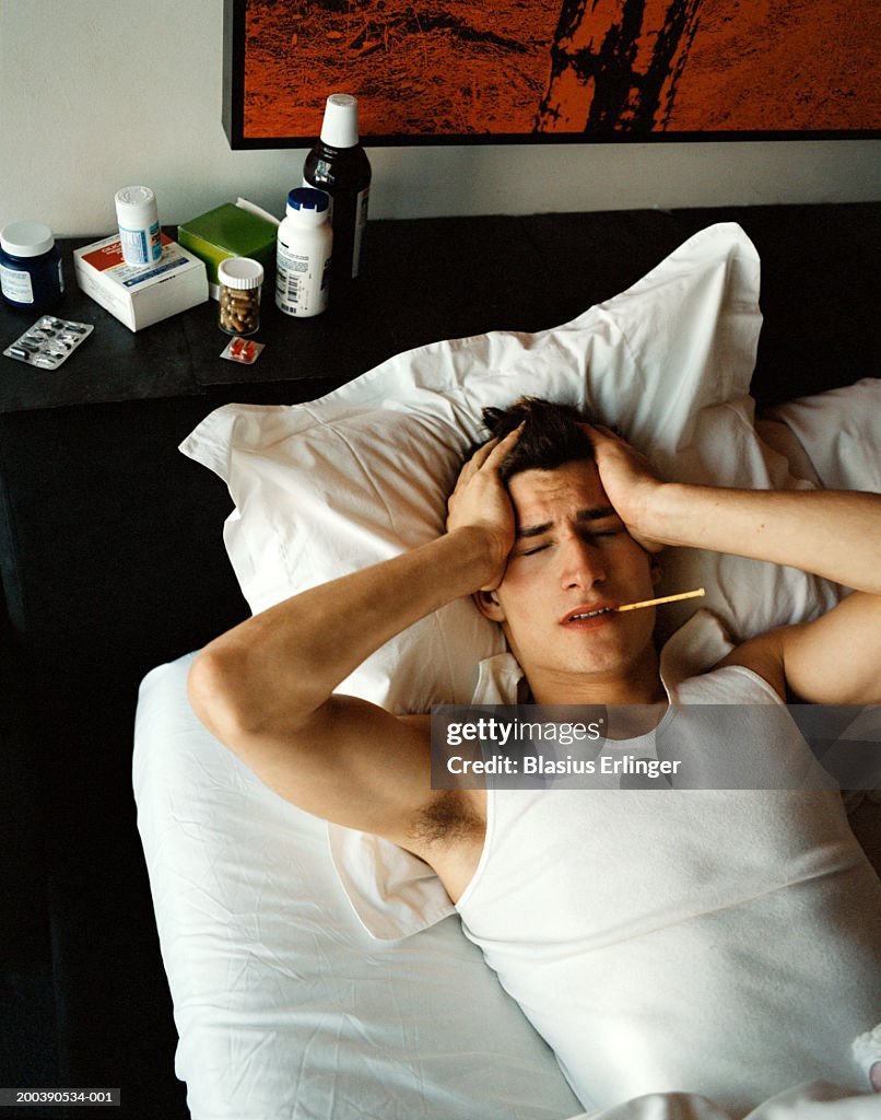 Young man in bed with thermometer in mouth holding head