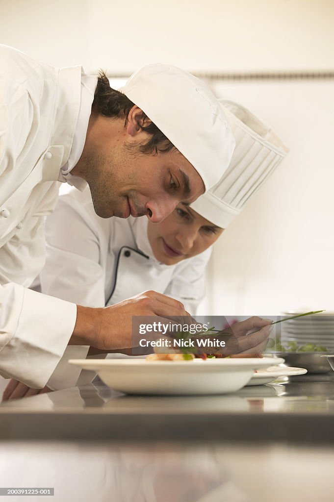 Female chef and male cook placing garnish on dishes, smiling