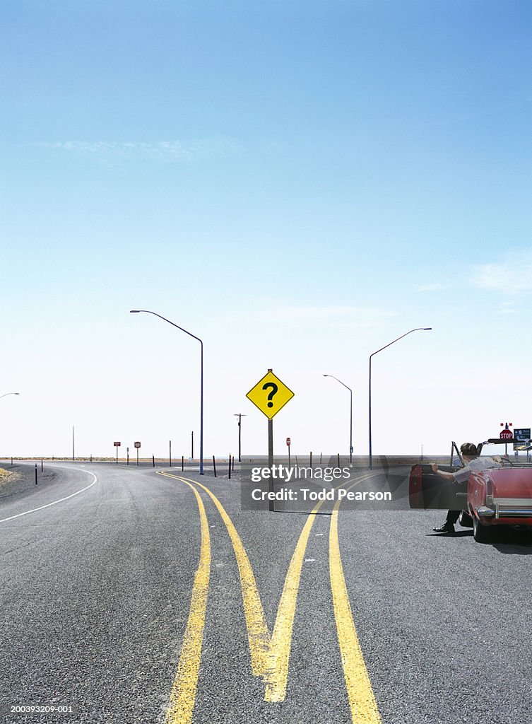 Man in convertable car at split in road, question mark traffic sign