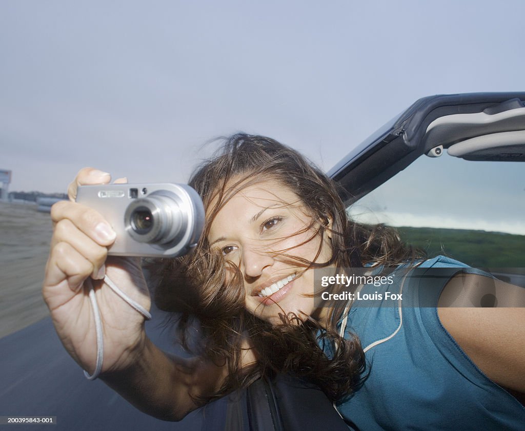 Young woman leaning out of convertible car, taking photograph