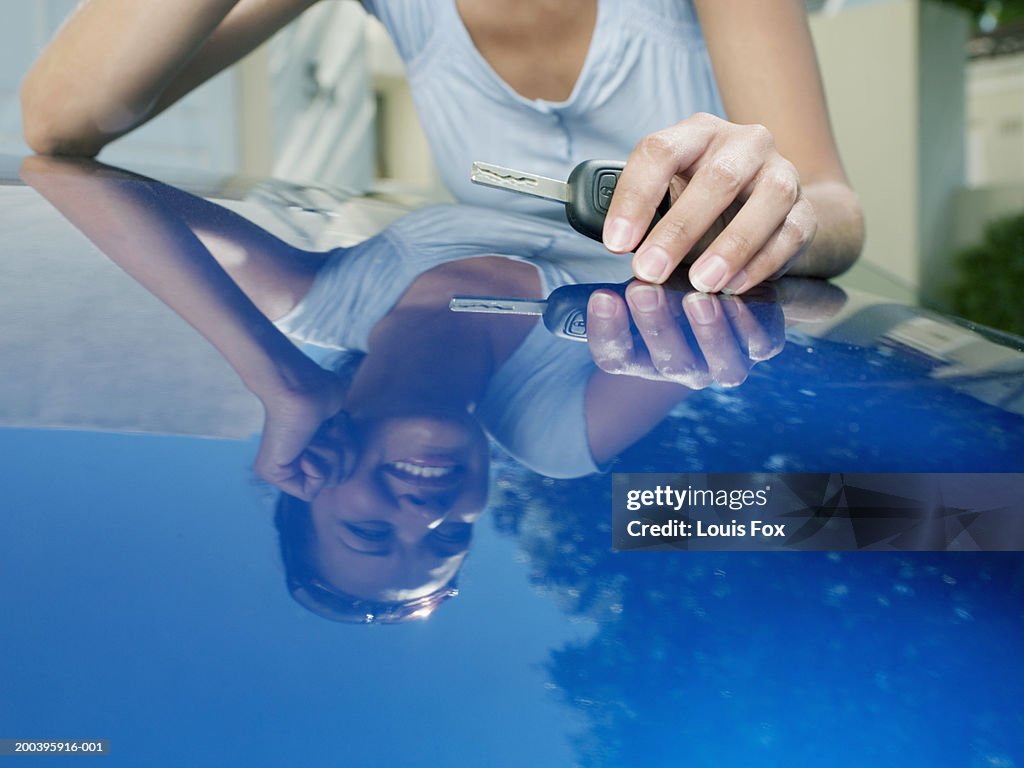 Young woman leaning on car, holding keys, reflection in car bonnet