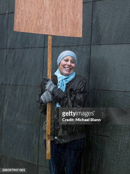 mature woman standing in rain holding placard, looking away, laughing - aktivist stock-fotos und bilder