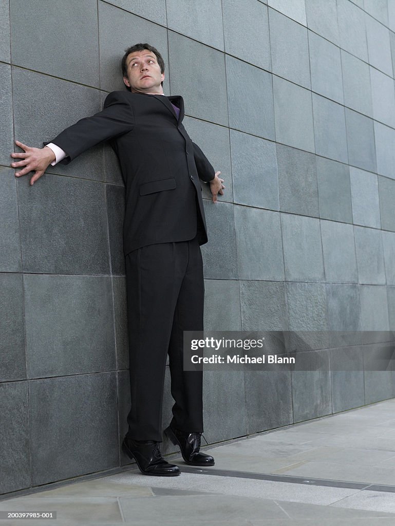 Businessman on tip toes, back against wall, arms outstretched, looking up