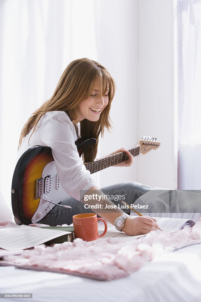 Teenage girl (14-16) with guitar writing in music book on bed, smiling