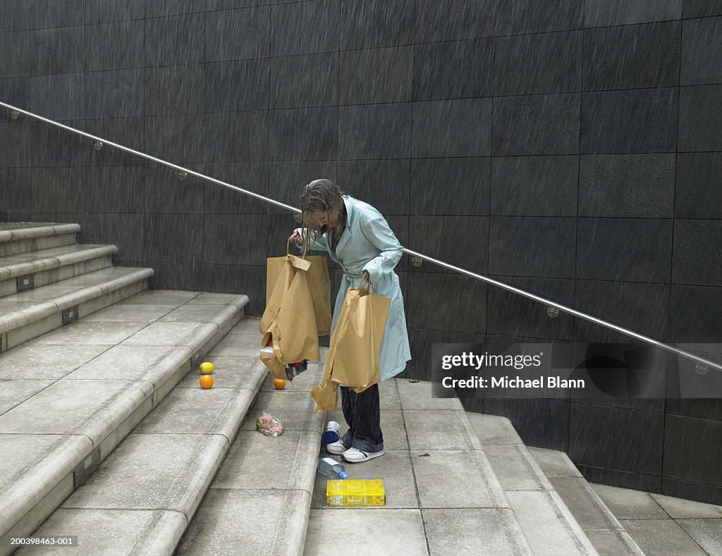 Mature woman in rain holding split bags, groceries spilling over steps