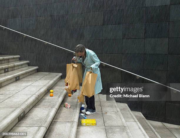 mature woman in rain holding split bags, groceries spilling over steps - carrying groceries stockfoto's en -beelden