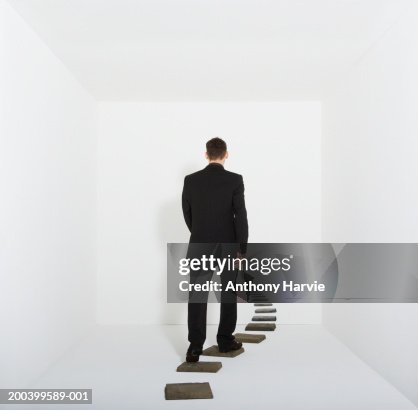 Businessman walking on stepping stones in white room, rear view Businessman walking on stepping stones in white room, rear view