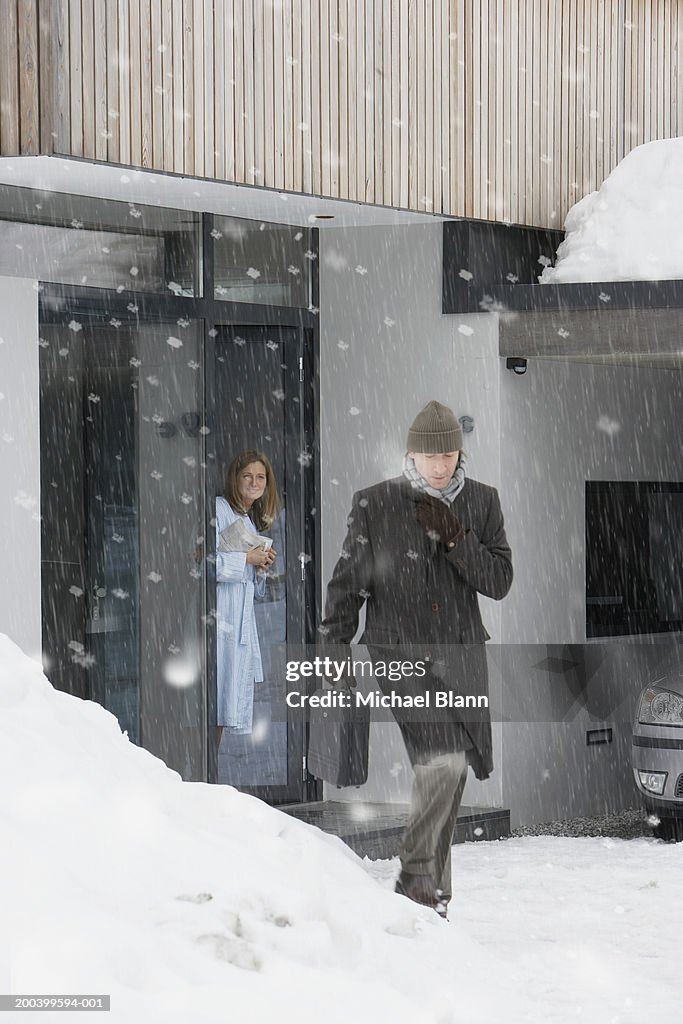 Man leaving house in snow, woman standing at front door, smiling