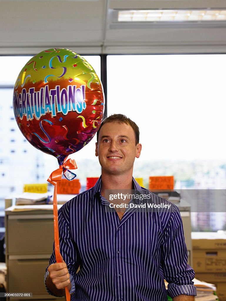 Young man holding 'congradulations' balloon in office, smiling