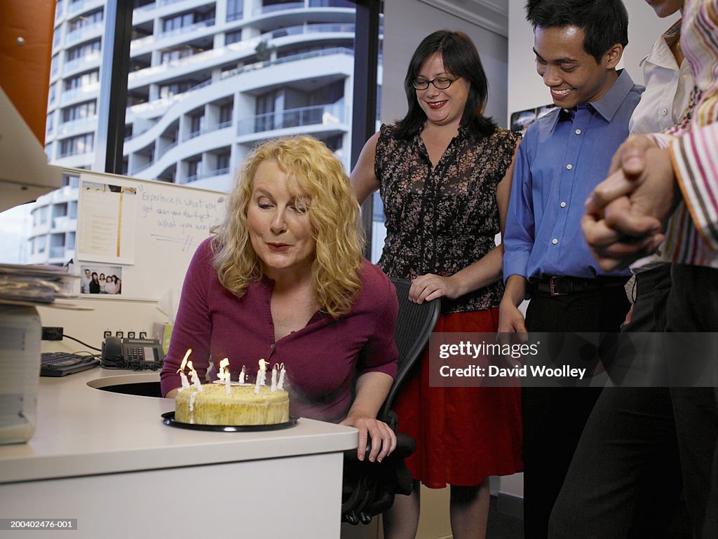 Office workers celebrating birthday, woman at desk blowing out candles on cake