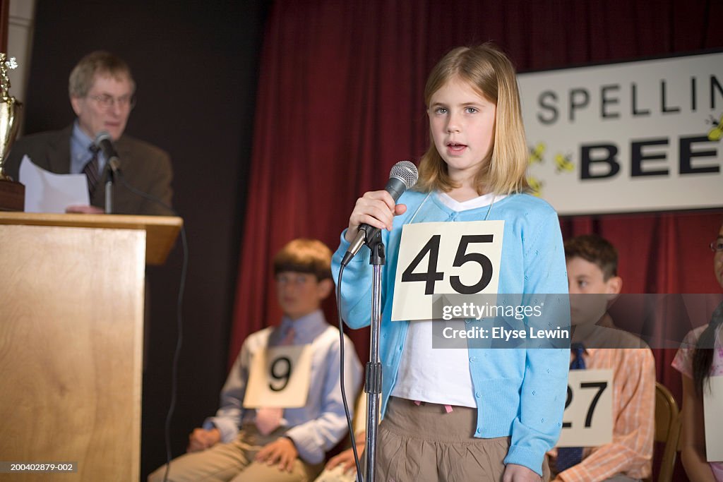 Girl (6-8) participating in spelling bee, standing at microphone