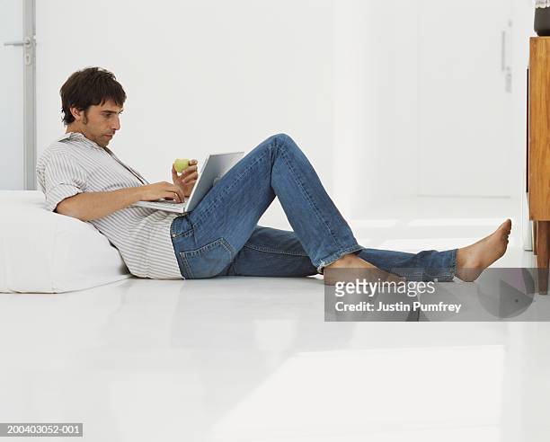 young man on floor leaning against mattress, using laptop, side view - jeans barefoot stock pictures, royalty-free photos & images