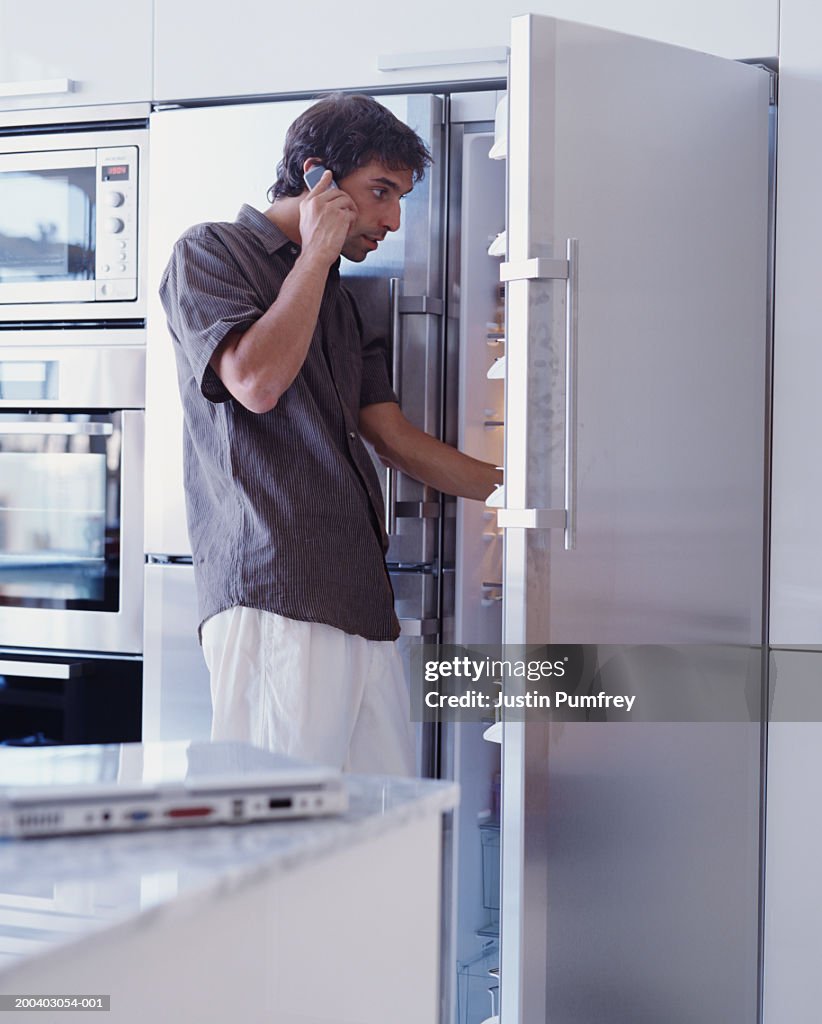 Young man looking in fridge whilst using mobile phone, side view