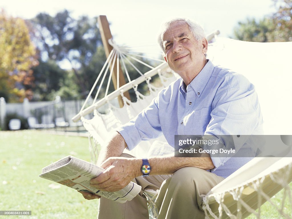 Senior man sitting on hammock holding newspaper, portrait