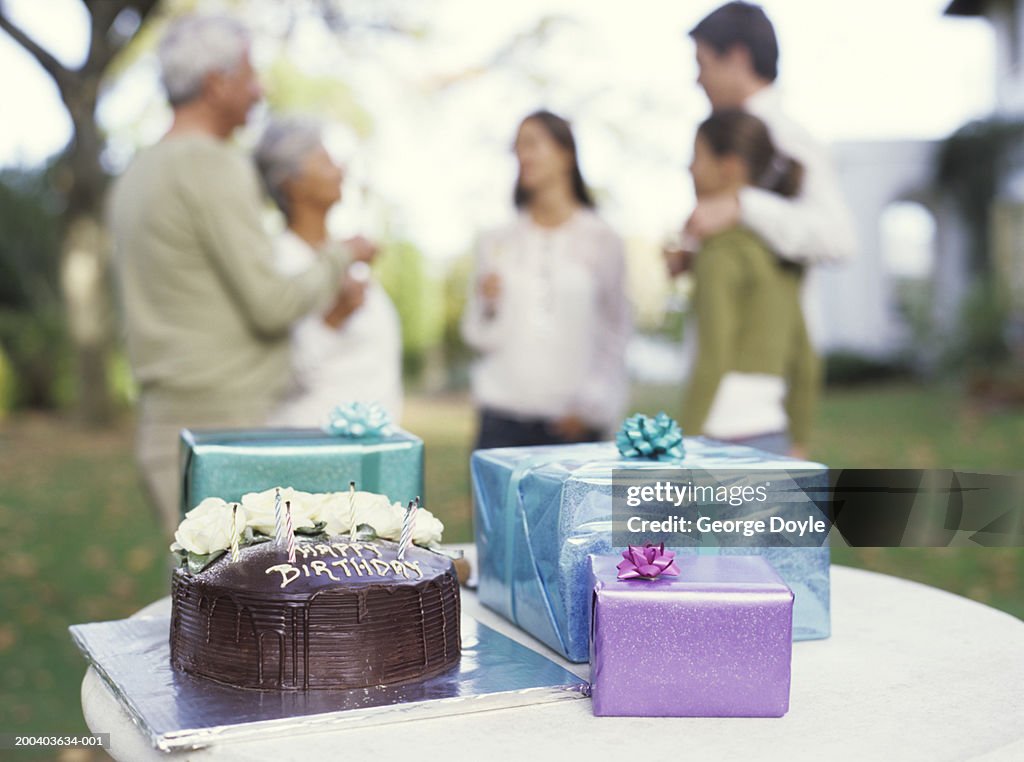 Gifts and birthday cake on table, close up, family in background