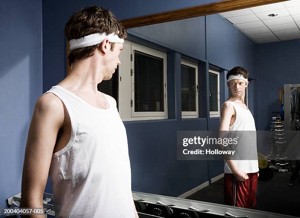 man wearing sweatband looking at reflection in gym mirror, flexing arm - skinny nerd stock pictures, royalty-free photos & images