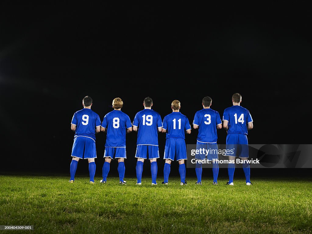 Six young male soccer players standing side by side, rear view
