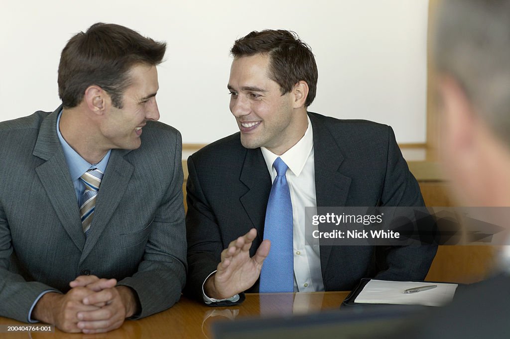 Two businessmen at table, one making hand gesture, smiling