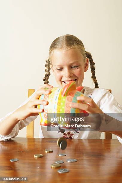 girl (10-12) emptying piggy bank onto table, smiling - blonde girl piggy bank stock-fotos und bilder