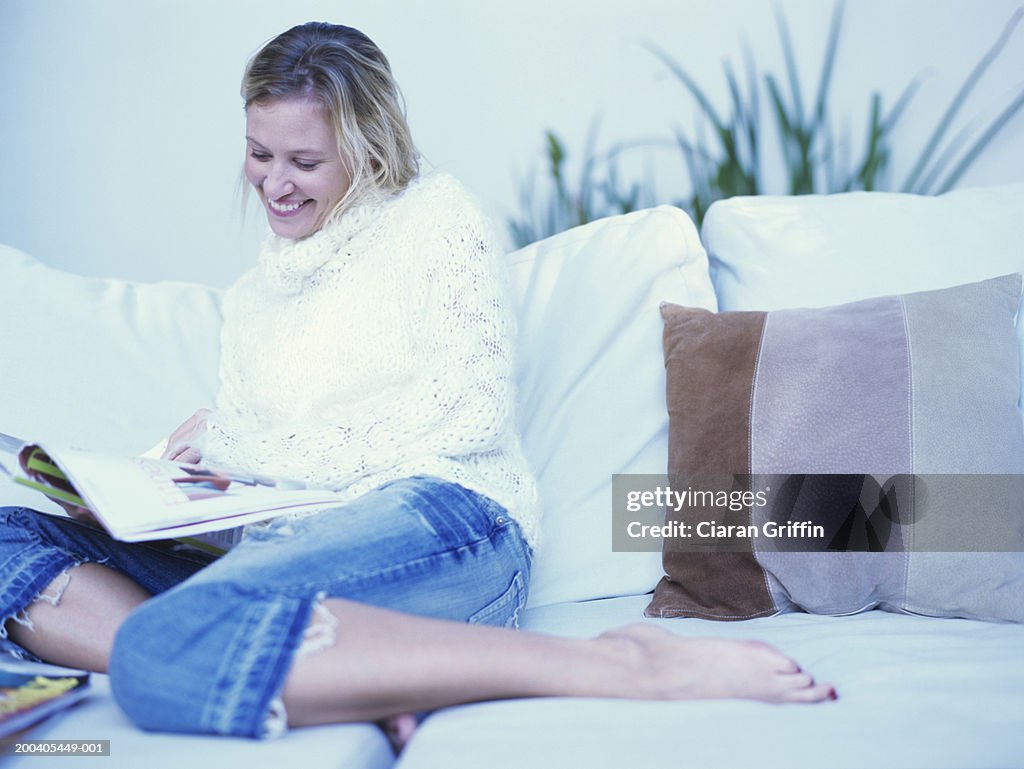 Young woman sitting on sofa reading magazine, smiling