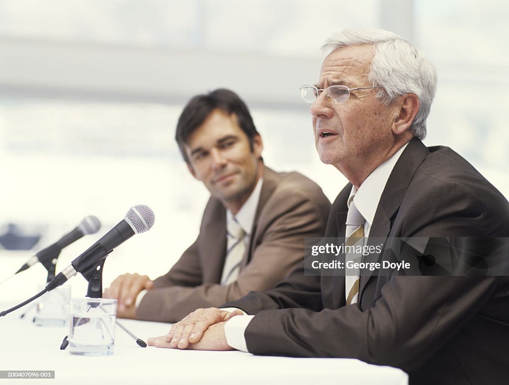 Two businessmen sitting by microphones, senior man speaking