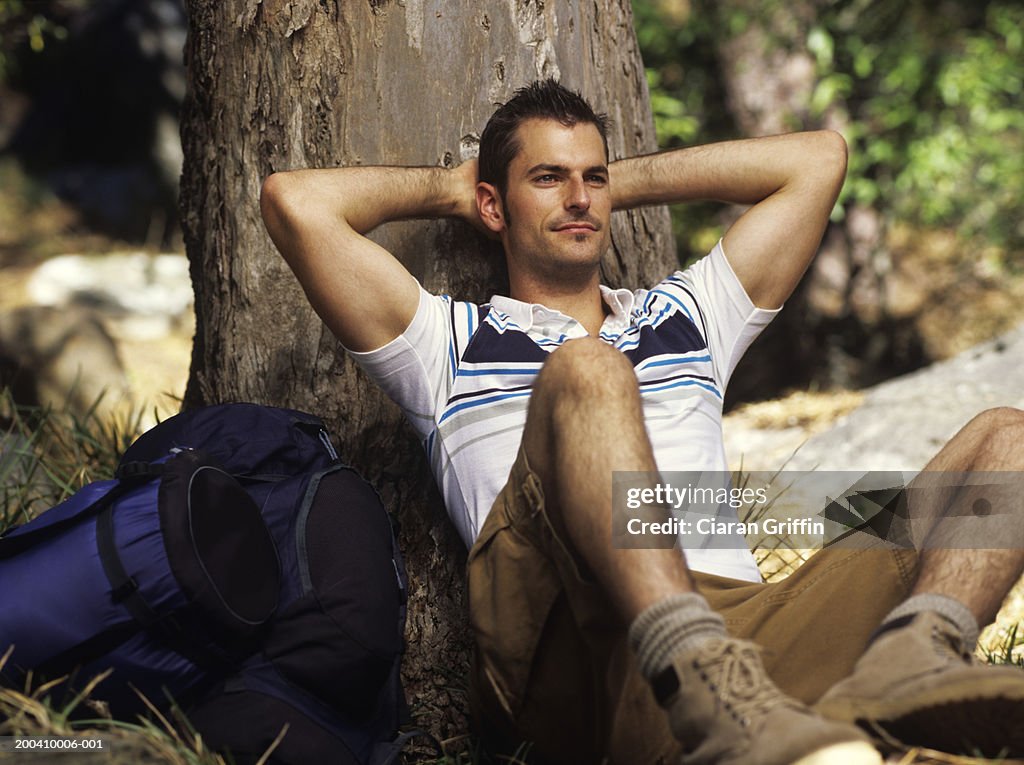 Young man with rucksack sitting against tree trunk