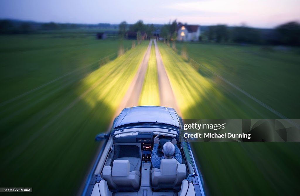 Mature man driving on rural road in convertible car with headlights on
