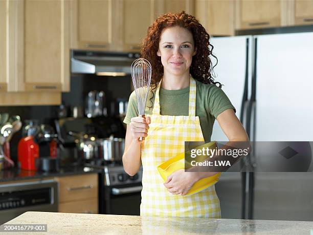 young woman in kitchen, holding wire wisk and bowl, portrait - ballonklopper stockfoto's en -beelden