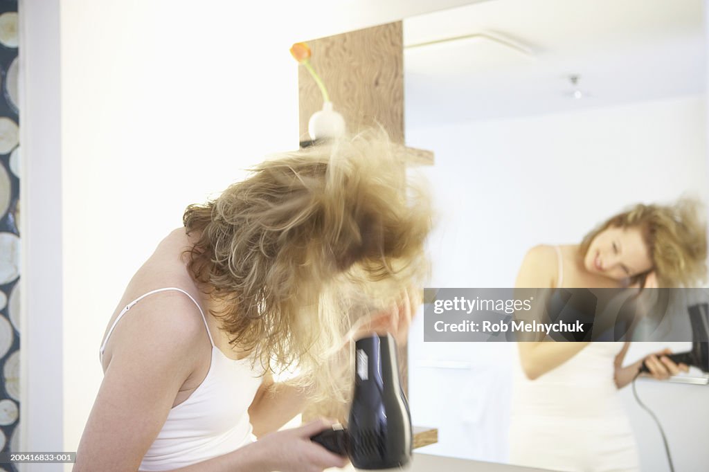 Young woman blow drying hair in bathroom, side view (blurred motion)