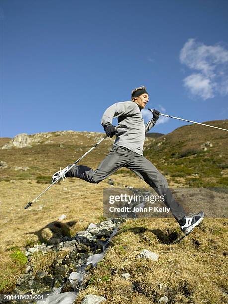 man nordic walking, leaping across stream, side view (wide angle) - hiking pole stockfoto's en -beelden