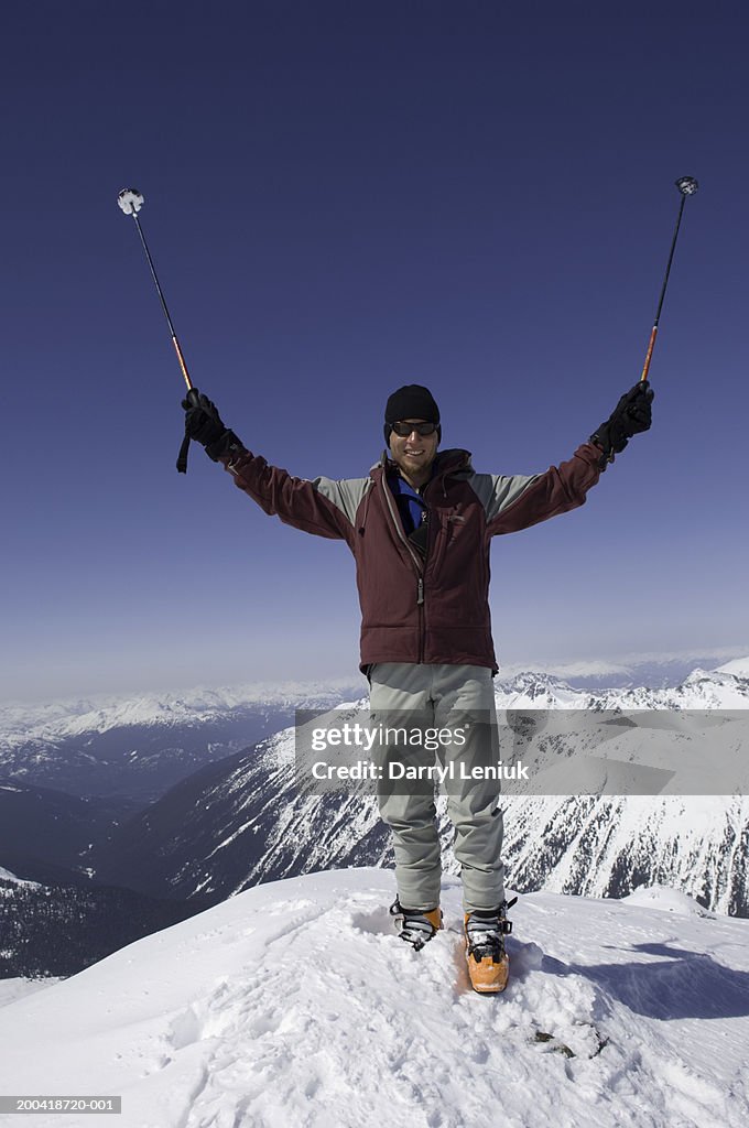 Male mountaineer with ski poles atop snowy peak, arms raised, portrait