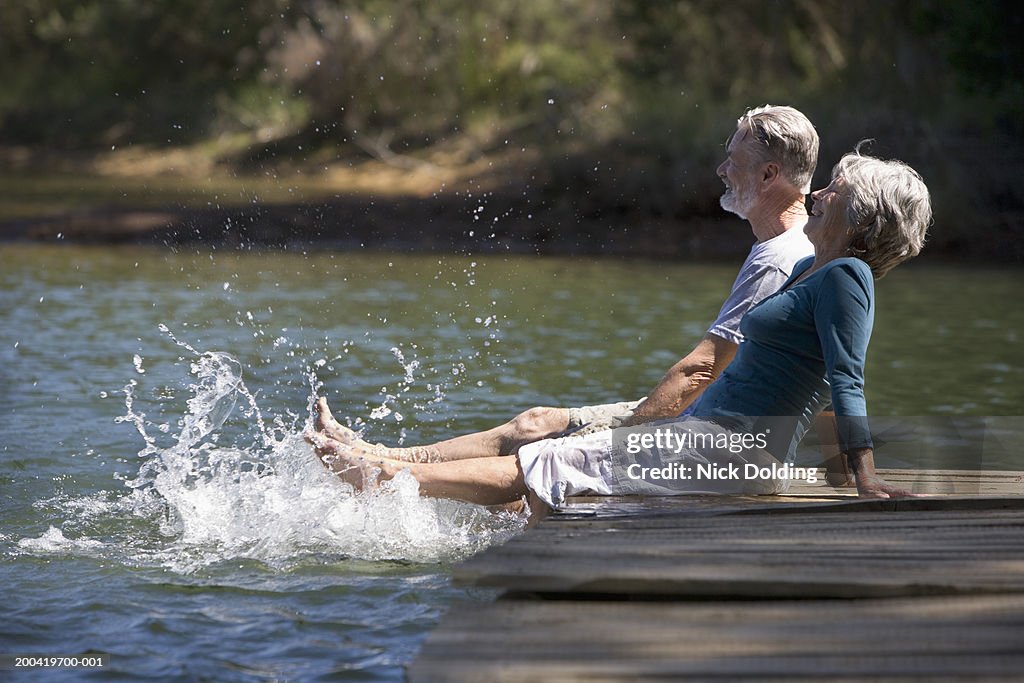 Senior couple sitting on lake jetty splashing feet in water, smiling