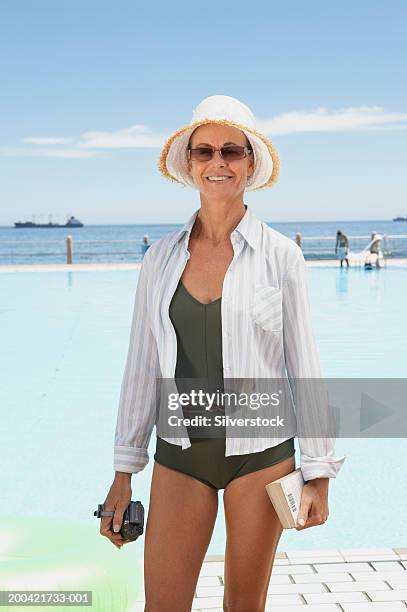 mature woman standing by pool, wearing sun hat and glasses, smiling - middle aged woman bathing suit stock pictures, royalty-free photos & images