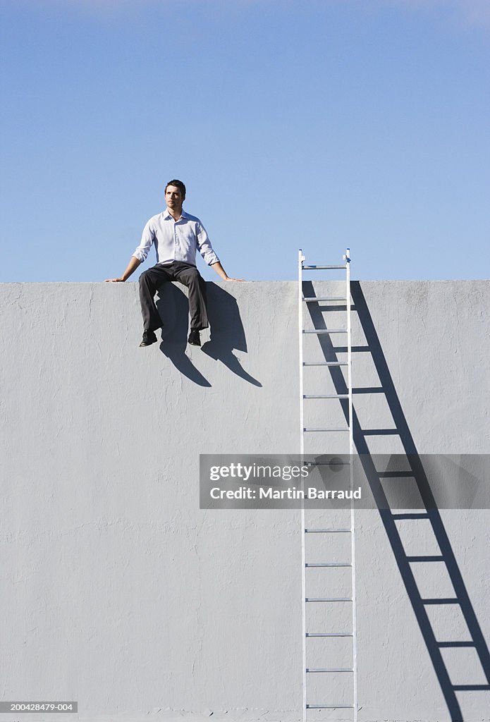 Businessman sitting on top of concrete wall, ladder against wall
