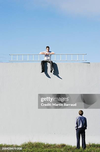 man, rear view, looking up at man sitting on wall holding ladder - malignity stockfoto's en -beelden