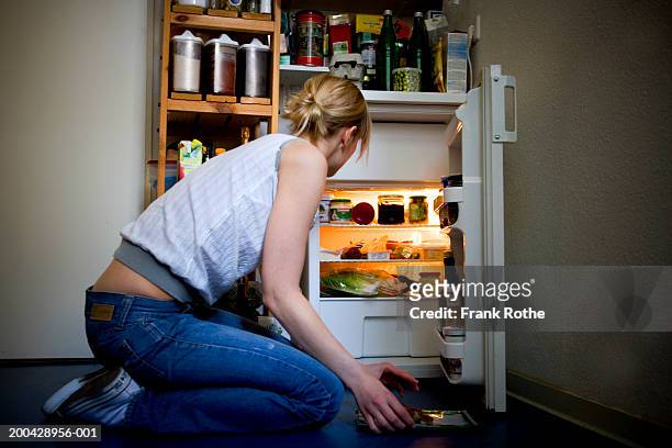young woman kneeling by open fridge in kitchen, side view - open fridge stock-fotos und bilder
