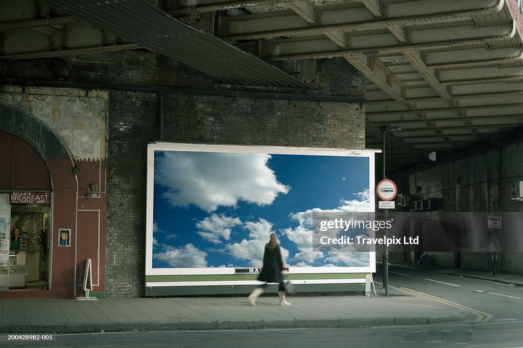 Woman walking past billboard poster of cloudy sky on city street