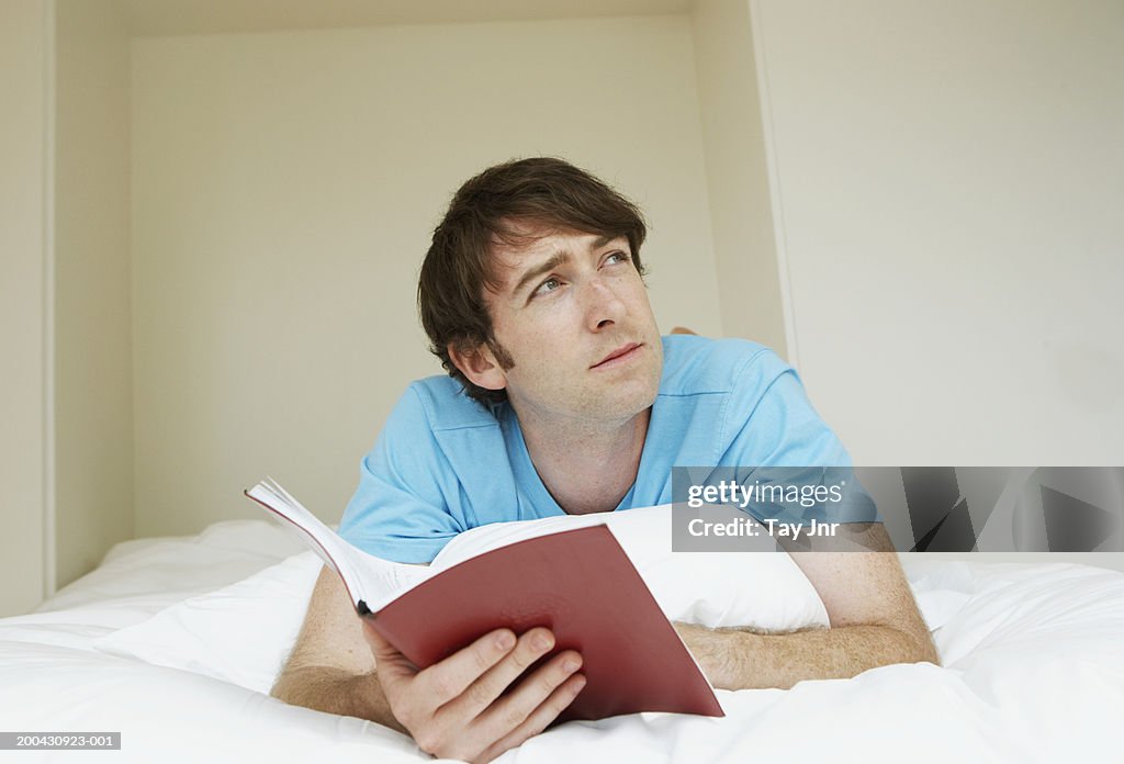 Young man lying on bed holding open book, looking up