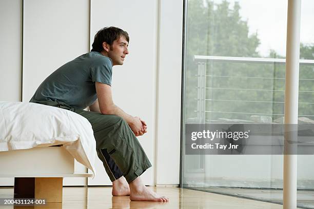 young man sitting on edge of bed, looking out patio doors, side view - men in bed 個照片及圖片檔