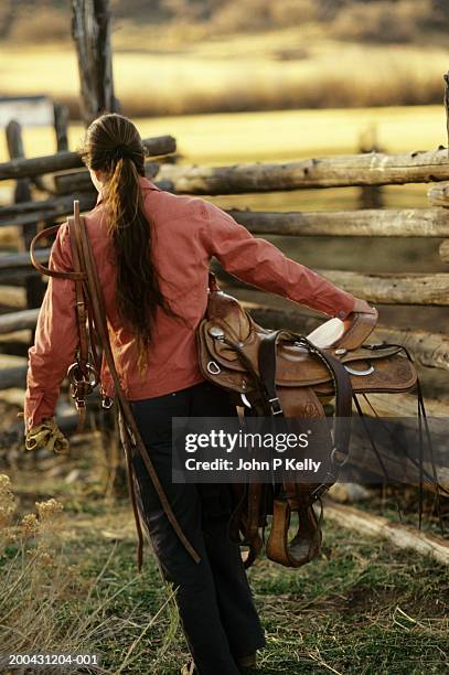 woman carrying horse tack, walking by corral, rear view - cowgirl hairstyles stock-fotos und bilder