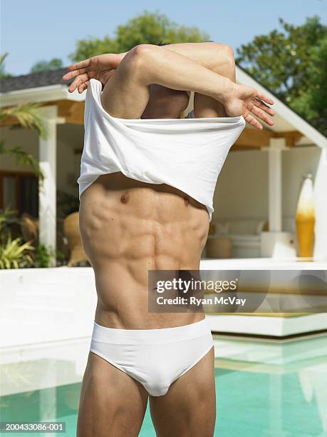 young man removing shirt beside swimming pool, arms raised - man wearing speedo stock-fotos und bilder