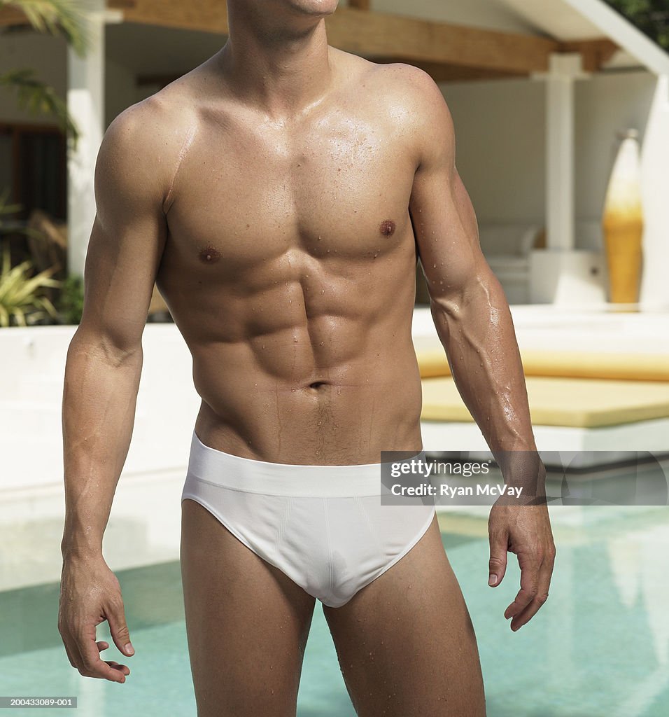 Young man in racing briefs standing beside pool, mid section