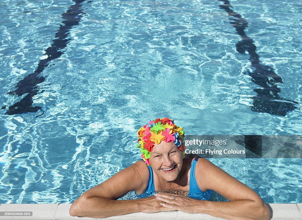 Senior woman wearing swimming hat, resting on edge of pool, smiling